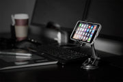 A smartphone held by the Mobile Grip 2 Phone Car Mount with Sticky Suction is mounted on a stand before a computer monitor, with a keyboard, mouse, stylus, and paper coffee cup on a dark desk in a modern workspace.