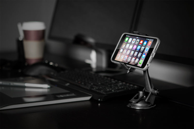 A smartphone held by the Mobile Grip 2 Phone Car Mount with Sticky Suction is mounted on a stand before a computer monitor, with a keyboard, mouse, stylus, and paper coffee cup on a dark desk in a modern workspace.