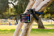 An 11 Adjustable Tripod Mount with Phone Holder is attached to a tree branch in a sunny park, ideal for content creators. Playground equipment and green grass appear in the background.