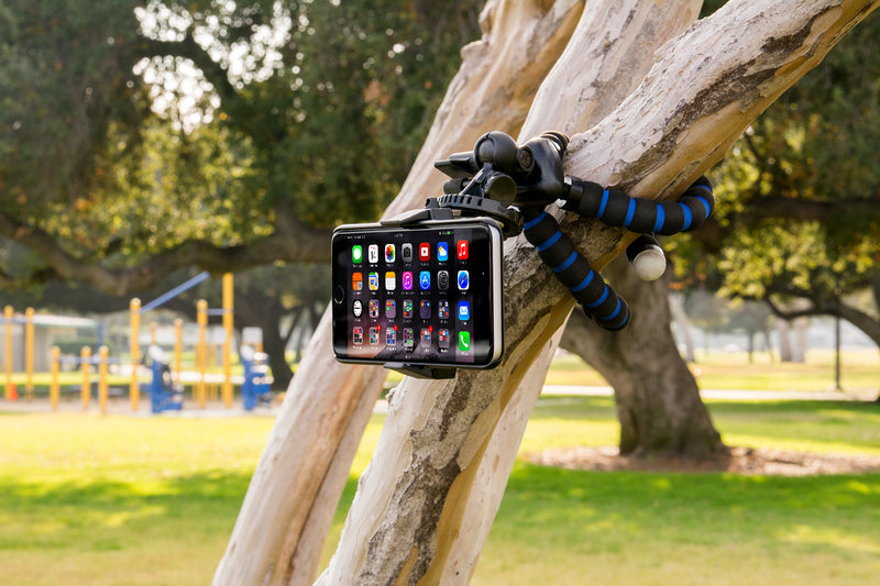 An 11 Adjustable Tripod Mount with Phone Holder is attached to a tree branch in a sunny park, ideal for content creators. Playground equipment and green grass appear in the background.