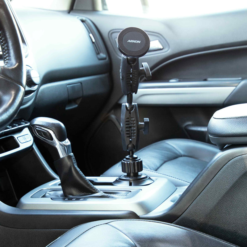 An Arkon Mounts Robust® Dual Shaft Cup Holder Mount with Magnetic Phone Holder (25mm, MagSafe compatible) is installed in the car’s center console cup holder next to the gear shift, with black leather seats and a modern dashboard in view.