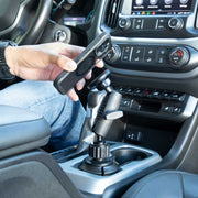 A person places a smartphone onto the Phone Cup Holder Mount with Magnetic Holder - MagSafe Compatible - 25mm, which is attached to the vehicle’s center console near the gear shift. Car dashboard controls and climate settings are visible.