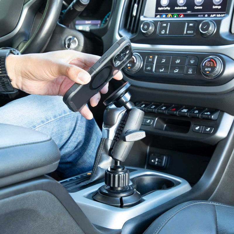 A person places a smartphone onto the Phone Cup Holder Mount with Magnetic Holder - MagSafe Compatible - 25mm, which is attached to the vehicle’s center console near the gear shift. Car dashboard controls and climate settings are visible.