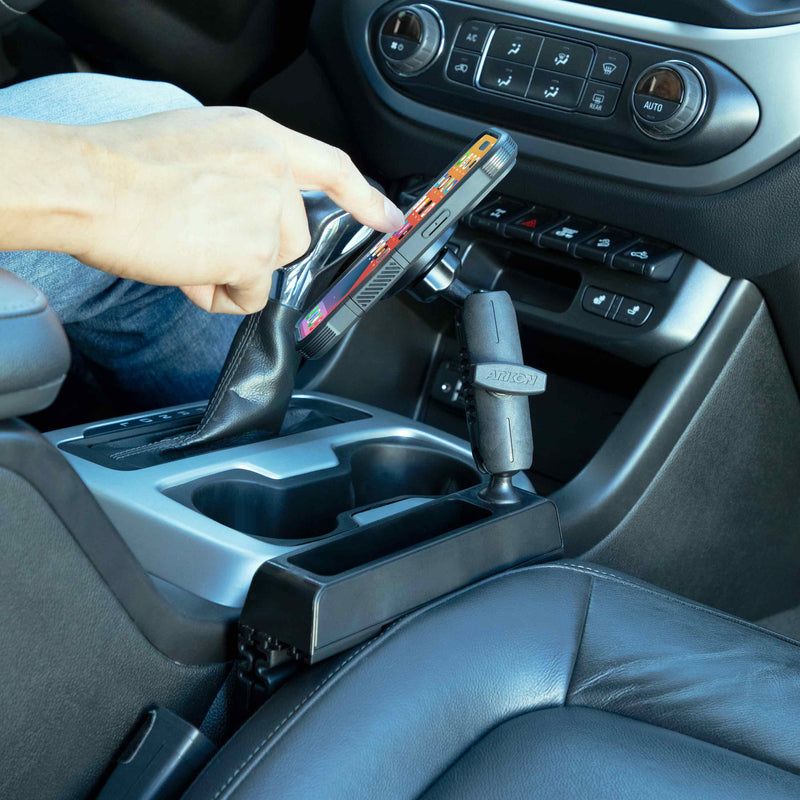 A hand shifts a car’s gear lever while holding a smartphone. The interior shows controls, cup holders, and a Magnetic Phone Holder Console Wedge Mount - MagSafe Compatible installed near the shifter.