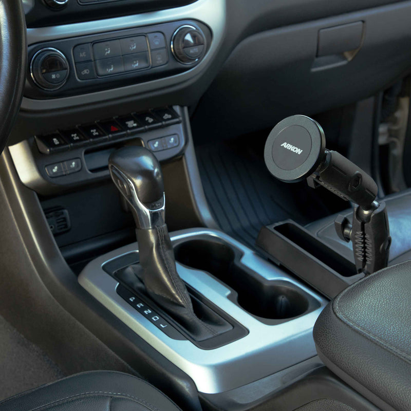 The car interior displays the center console featuring an automatic gear shift, cup holders, dashboard controls, and a Magnetic Holder with Seat Wedge Mount and Dual Shaft Arm—MagSafe Compatible—mounted on a flexible arm for convenient hands-free iPhone use. The floor mat is also visible.