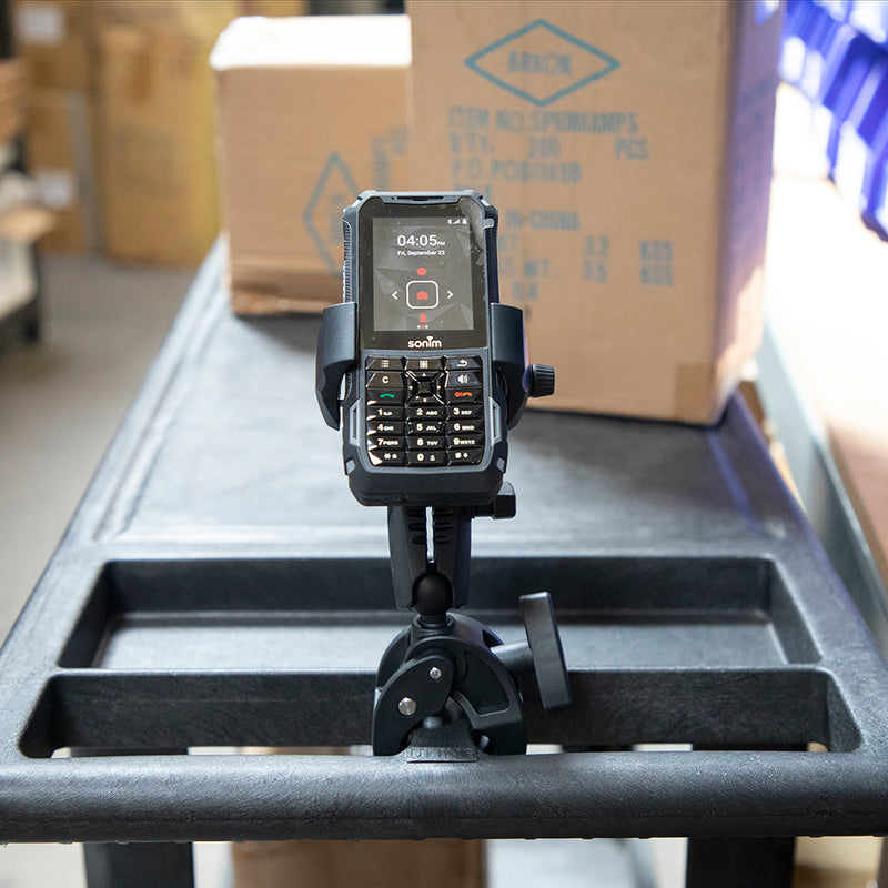 A black handheld device with a physical keypad is mounted in the RoadVise® Ultra Holder with Clamp Mount on a rolling cart in a warehouse, surrounded by boxes and shelves.