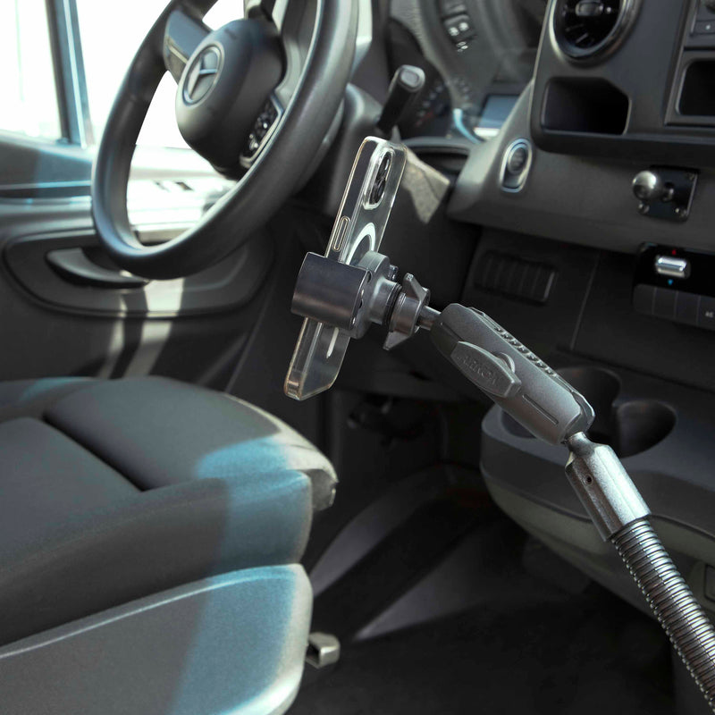 A smartphone is mounted on the RoadVise® Heavy-Duty Seat Rail or Floor Phone Mount near the driver’s seat and steering wheel, with the car’s interior and dashboard controls visible in the background.