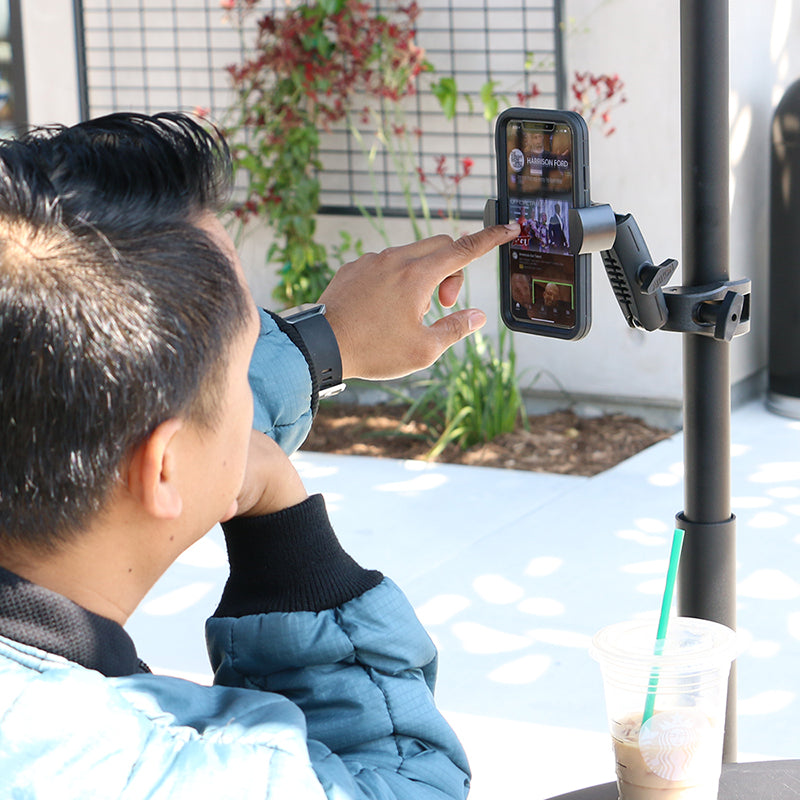A person at an outdoor table uses the RoadVise® Robust Clamp Phone Mount with Security Knob to secure their smartphone on a pole. An iced drink with a green straw is nearby, with plants and a metal grid in the background.