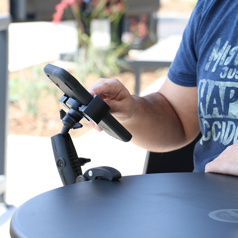 A person in a blue t-shirt uses their smartphone secured in a RoadVise® Robust Clamp Phone Mount with Security Knob, attached to the edge of an outdoor round black table.