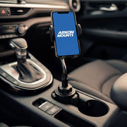 A Cup Holder Phone Mount with Gooseneck and 17mm Ball is installed in the car’s cup holder, holding a smartphone that displays the Arkon Mounts logo. The image highlights the vehicle’s gear shift and interior features.