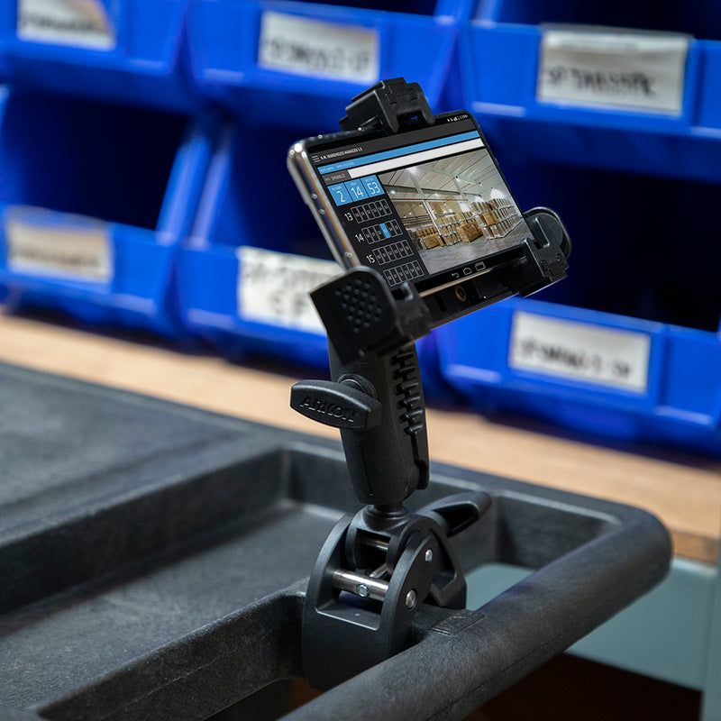 A smartphone is mounted on the LockVise™ Phone Clamp Mount with 3.75” Shaft Arm, which is attached to a cart and displaying a building interior. Blue storage bins with labels are visible in the background.