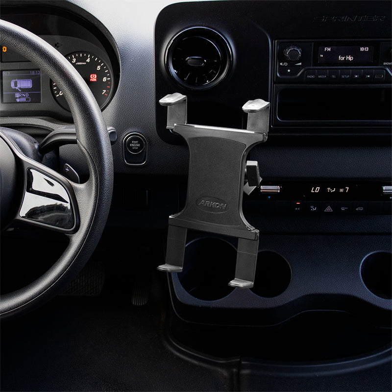 A black car interior with a steering wheel, dashboard, and an empty Slim-Grip® Tablet Holder with Double Socket Swivel Arm and Drill-Base Mount installed near the center console. The radio, air controls, and cup holders are also visible.