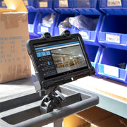 A rugged tablet, securely held by the LockVise™ Locking Tablet Clamp Mount with 38mm Ball Compatibility, displays a warehouse inventory interface while mounted on a cart in an industrial warehouse, with blue storage bins and cardboard boxes in the background.
