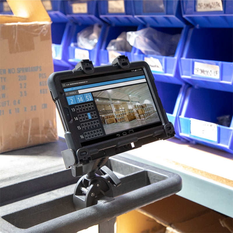 A rugged tablet, securely held by the LockVise™ Locking Tablet Clamp Mount with 38mm Ball Compatibility, displays a warehouse inventory interface while mounted on a cart in an industrial warehouse, with blue storage bins and cardboard boxes in the background.