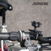 Close-up of a bicycle handlebar with the Bike or Motorcycle Handlebar Mounting Pedestal - Dual-T Compatible attached. Part of the bike seat and blurred pavement are visible in the background, with the Arkon logo in the upper right corner.