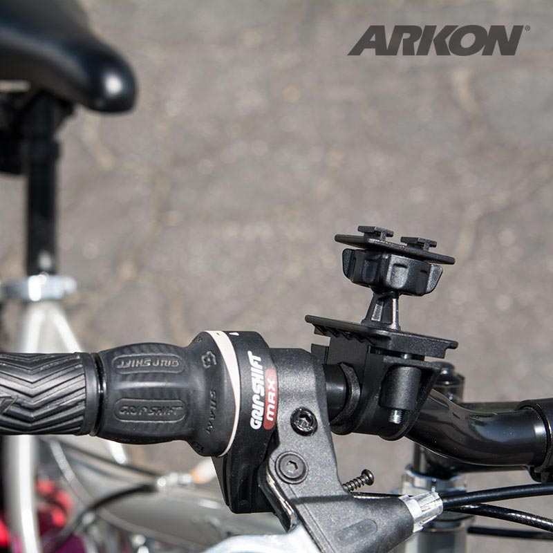 Close-up of a bicycle handlebar with the Bike or Motorcycle Handlebar Mounting Pedestal - Dual-T Compatible attached. Part of the bike seat and blurred pavement are visible in the background, with the Arkon logo in the upper right corner.