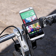 An iPhone is secured to a bike handlebar with the Bike or Motorcycle Mega Grip™ Phone Holder Strap Mount. The phone displays time, weather, and a video, connected by cable, while the bike stands on a paved outdoor surface.