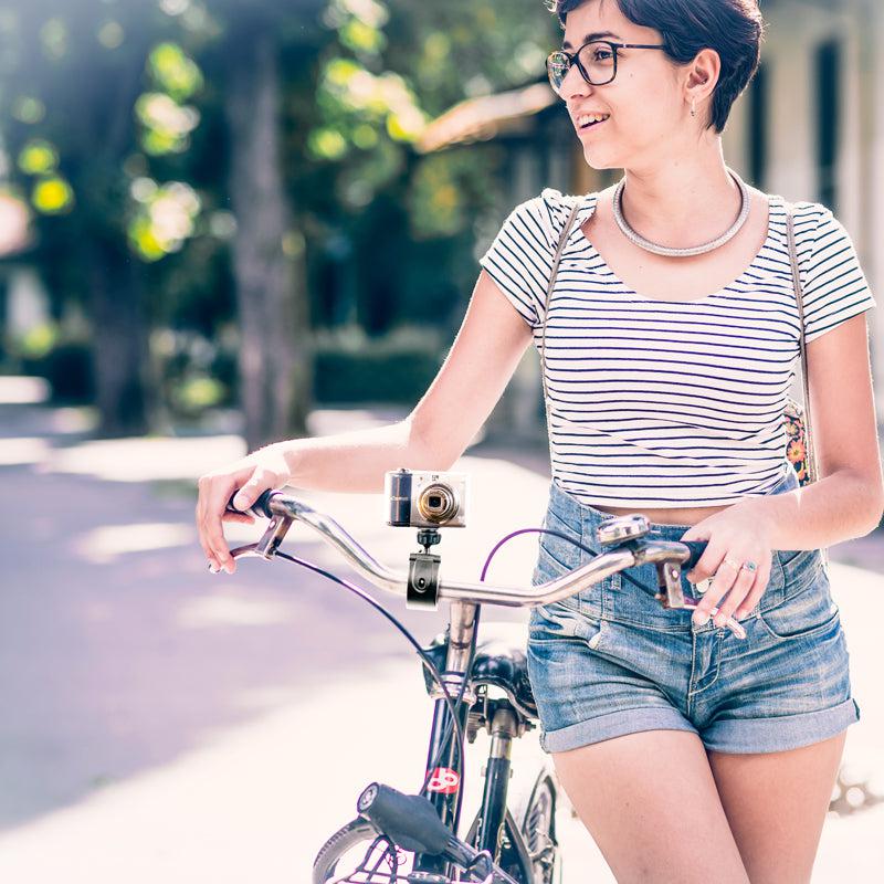 A woman in glasses, a striped shirt, and denim shorts stands outdoors smiling, holding her bike equipped with the Handlebar Clamp Camera Mount. Trees are visible in the background.