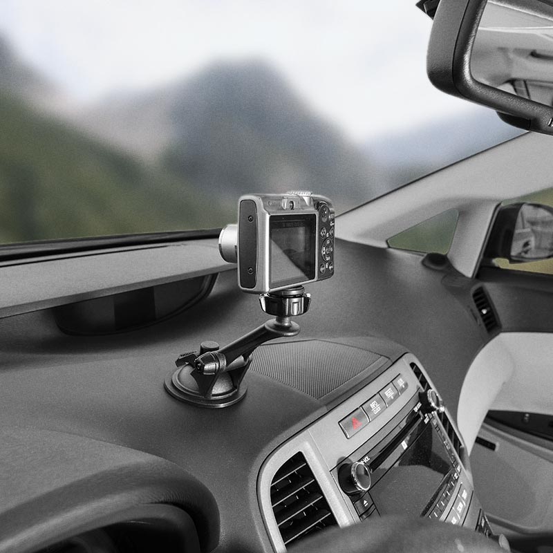 A Camera Holder with Sticky Suction Mount and Adhesive Disk is attached to a car dashboard, securing a camera aimed at the windshield with blurred mountains visible in the background.