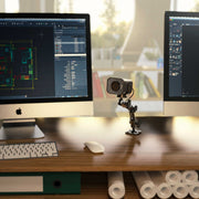 A small robot with a Camera Wall Mount with Multi-Angle Arm for Security Camcorders & Cameras as its head stands on a wooden desk between two monitors with design software, surrounded by rolled-up papers and office supplies.