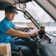 A man in a blue shirt and cap sits in a vehicle, reaching to adjust his smartphone on the Drill-Base Mega Grip™ Phone Mount for iPhone, Galaxy, Note, and more, while a package rests on the passenger seat.