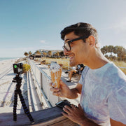 A man wearing glasses smiles while holding an ice cream cone at a sunny beach. A Flexible 11 Tripod for GoPro HERO Action Cameras is set up on the railing next to him, with palm trees, houses, and people in the background.