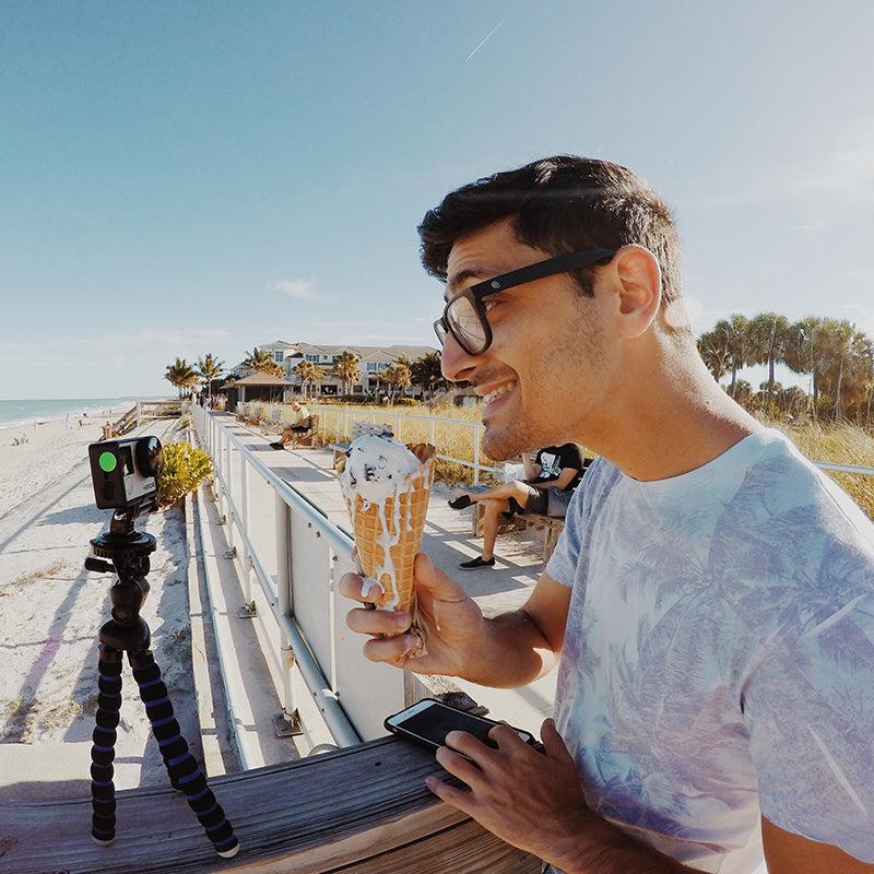 A man wearing glasses smiles while holding an ice cream cone at a sunny beach. A Flexible 11 Tripod for GoPro HERO Action Cameras is set up on the railing next to him, with palm trees, houses, and people in the background.