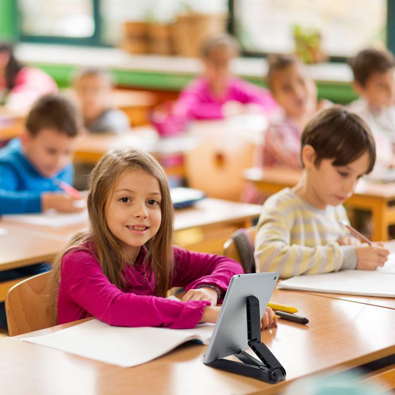 A young girl in a pink shirt smiles at her classroom desk, using her tablet on the Arkon Mounts Portable Tablet & Phone Stand, with an open notebook. Other children work in the background.