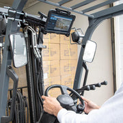 A person operates a forklift in a warehouse, where a LockVise™ Locking Forklift Front Guard Tablet Mount with 10.25 inch Metal Robust™ Shaft secures a tablet above the dashboard showing inventory. Stacks of cardboard boxes are seen in the background.