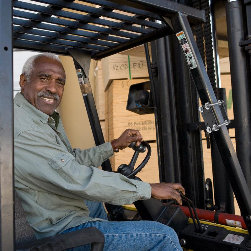 An older man smiles while operating a forklift equipped with the Forklift Front Guard Mount in a warehouse, with stacked wooden crates in the background.
