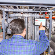 A man in a blue plaid shirt operates a forklift labeled 5000 lbs capacity in an industrial setting, using a touchscreen device secured by the Forklift Slim-Grip® Tablet Mount.