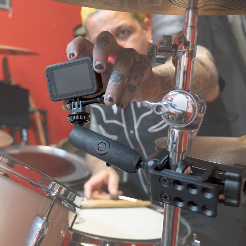 A tattooed drummer fixes a Heavy-Duty Adjustable Clamp Mount for GoPro HERO Action Cameras to a drum kit, holding drumsticks with multiple drums and cymbals in view—an essential accessory for live music streaming.