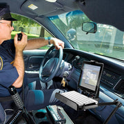 A police officer sits in a patrol car, speaking into a radio mic. A Heavy-Duty Car Seat Rail Tablet Mount with Keyboard Tray Combo holds a laptop displaying official documents in front of the dashboard. Keys hang from the ignition.