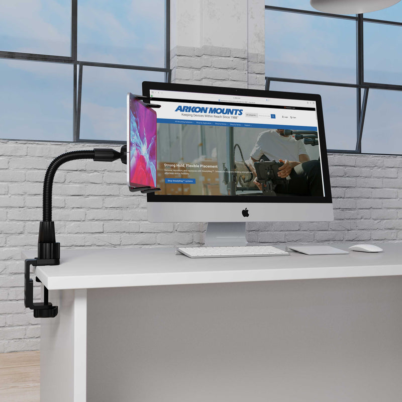The Heavy-Duty Slim-Grip® Tablet Clamp Mount secures a smartphone to a white desk next to an iMac showing the Arkon Mounts website in a modern office with large windows and a white brick wall.