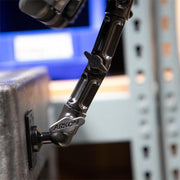 Close-up of the Slim-Grip® Tablet Holder with Drill-Base Mount and 8 Multi-Angle Arm attached to a desk edge, with a metal shelving unit and blue screen blurred in the background.