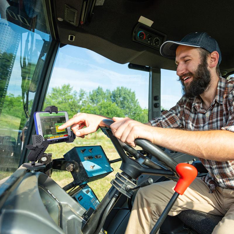A bearded man in a plaid shirt and cap smiles while using a touchscreen device on the LockVise™ Locking Phone Mount with 2.75 Arm and Metal AMPS Drill Base, installed near the tractors steering wheel. Greenery and blue sky are visible outside.