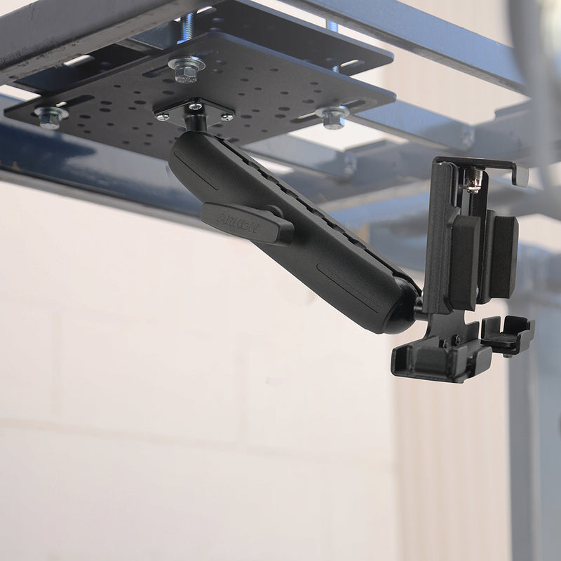 A LockVise™ Metal Locking Overhead Guard Tablet Mount with an 8.5″ Metal Robust™ Shaft is attached to a metal frame, functioning as a sturdy tablet holder and securely supporting a downward-facing device clamp. The background is out of focus, featuring a light-colored wall.