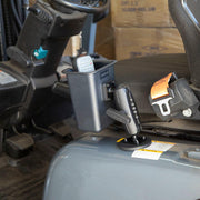 A Magnetic Mount with Barcode Scanner Holster Holder secures a handheld device to a vehicle seat’s cup holder, with a seatbelt visible and stacked boxes in the background.