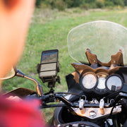 Close-up of a person riding a motorcycle, with the Mega Grip 25mm Robust Aluminum Motorcycle Handlebar Smartphone Mount holding a smartphone running navigation; grassy field and trees softly blurred in the background.