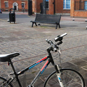 A black and blue bicycle with a Mega Grip 25mm Robust Aluminum Motorcycle Handlebar Smartphone Mount is parked on a paved plaza with a bench, trash can, and brick buildings. The image highlights the bikes handlebars and front wheel.