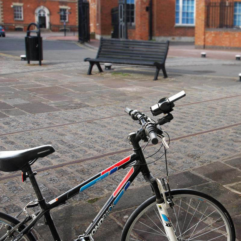 A black and blue bicycle with a Mega Grip 25mm Robust Aluminum Motorcycle Handlebar Smartphone Mount is parked on a paved plaza with a bench, trash can, and brick buildings. The image highlights the bikes handlebars and front wheel.
