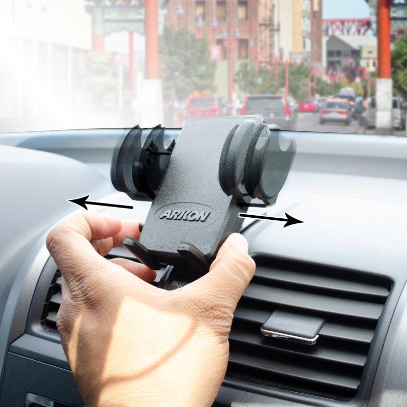 A hand adjusts the Mega Grip™ Car Phone Holder with Adhesive Mount for iPhone, Galaxy, and Note on a car’s air vent; arrows highlight the adjustable grips. The city view through the windshield shows hands-free calls are easy on the go.