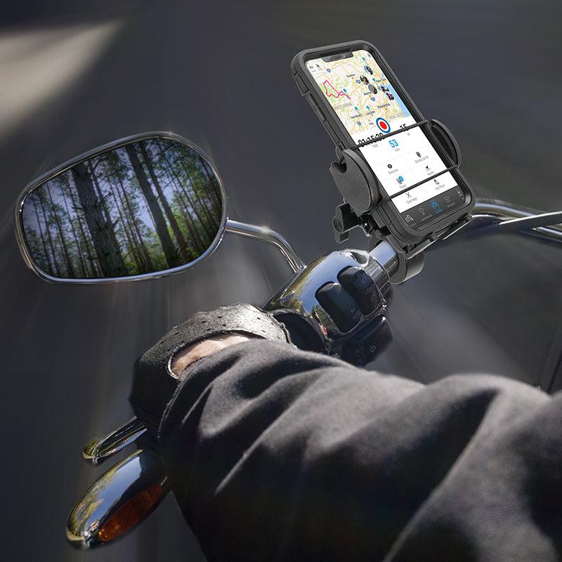A person wearing a black glove rides a motorcycle with a Mega Grip™ Premium Aluminum Motorcycle Handlebar Smartphone Mount securing a GPS smartphone to the handlebars. The side mirror reflects tall trees beneath a bright sky.