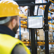 A warehouse worker in a yellow hard hat and vest checks inventory on a LockVise™ Metal Locking Forklift Front Guard Tablet Mount with 3.75 Shaft, with shelves of boxes visible in the background.