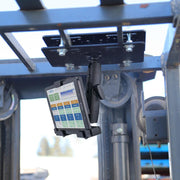 A tablet is mounted upside down on a LockVise™ Metal Locking Forklift Overhead Guard Tablet Mount with 6 Shaft attached to industrial equipment, showing a colorful interface. The background features a clear sky and blurred outdoor elements.