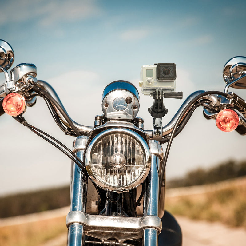 Close-up of a Premium Aluminum Motorcycle Handlebar Mount for GoPro Action Cameras attached near the handlebars and headlight, with a protective case, set against a blurred outdoor background under a blue sky.
