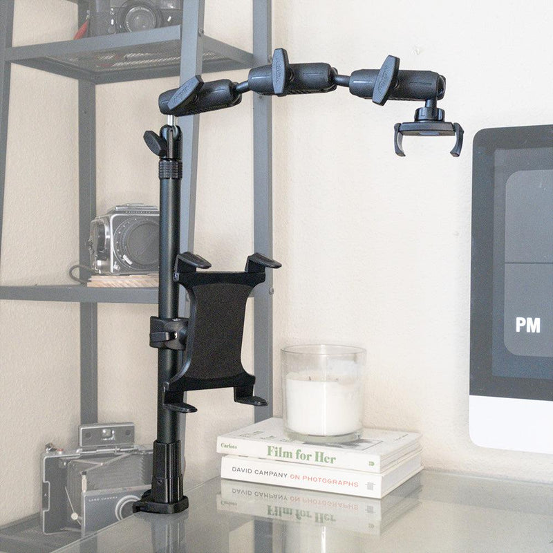 The Remarkable Creator™ Pro+Plus with Clamp Mount, in black, is attached to a glass desk next to a monitor, surrounded by a candle and stacked books, with vintage cameras displayed on a shelf in the background.