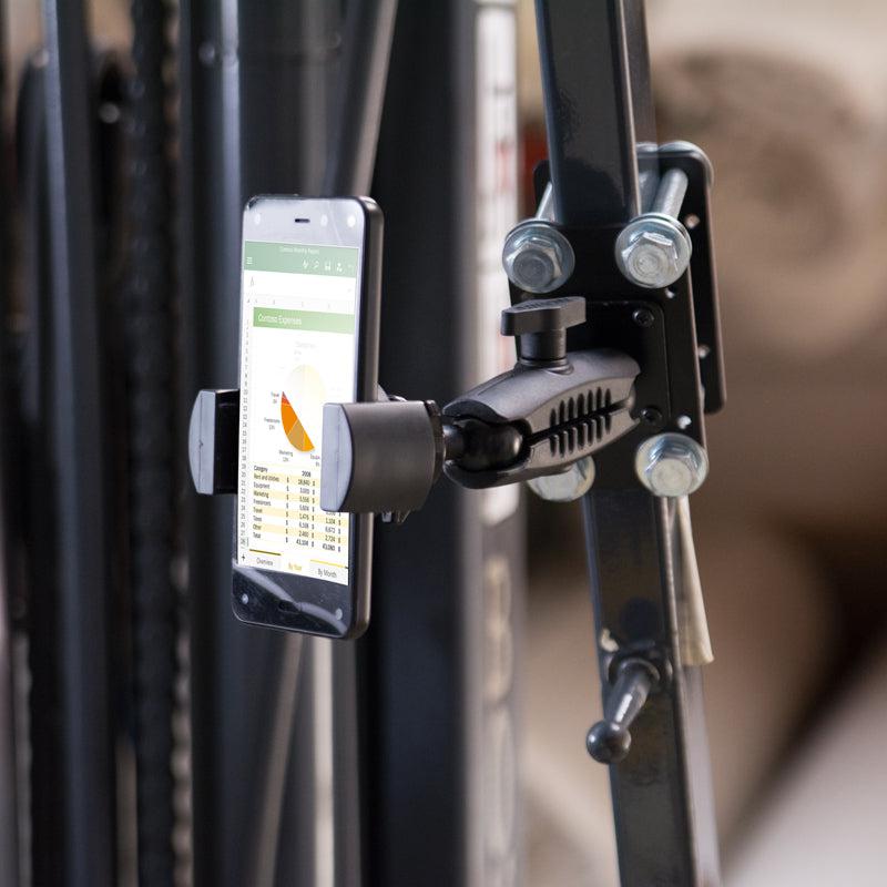 A smartphone showing a spreadsheet with a pie chart is held in a RoadVise® Forklift Front Guard Phone Mount attached to metal gym equipment, with a blurred background.