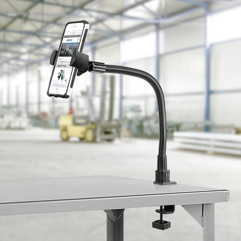 The RoadVise® Heavy-Duty Phone Clamp Mount with a 22 gooseneck is clamped to the edge of a gray table, firmly holding a smartphone; in the background, there’s a spacious, blurred industrial warehouse.