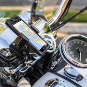 A RoadVise® Holder with Handlebar Mount secures a smartphone to motorcycle handlebars beside the speedometer, with blurred greenery and a street visible in the background.